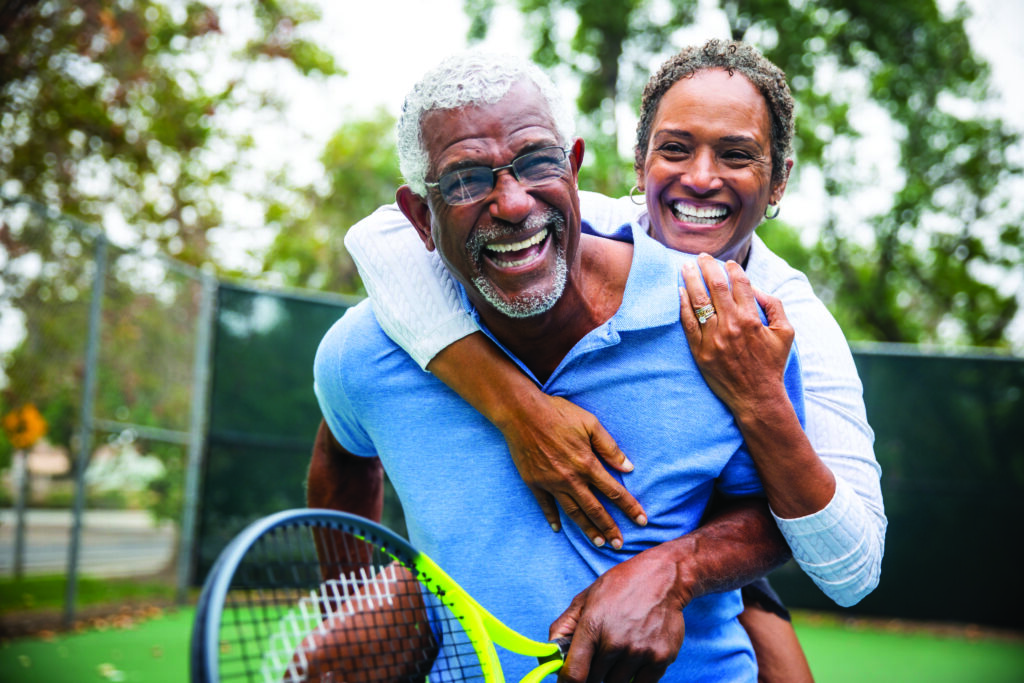 A senior black couple piggyback together on the tennis court.